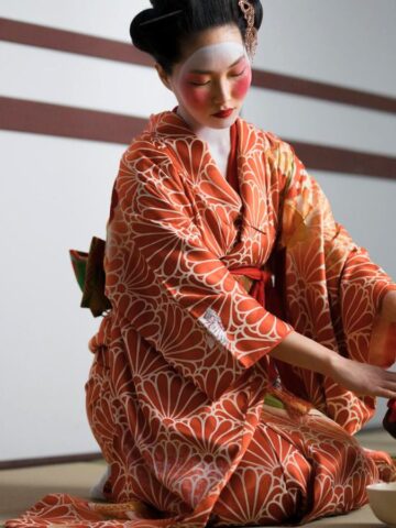 A person dressed in a traditional Japanese kimono is performing a tea ceremony. They are kneeling on a tatami mat and carefully pouring tea from a teapot into a cup. The background features a wall with wooden panels.