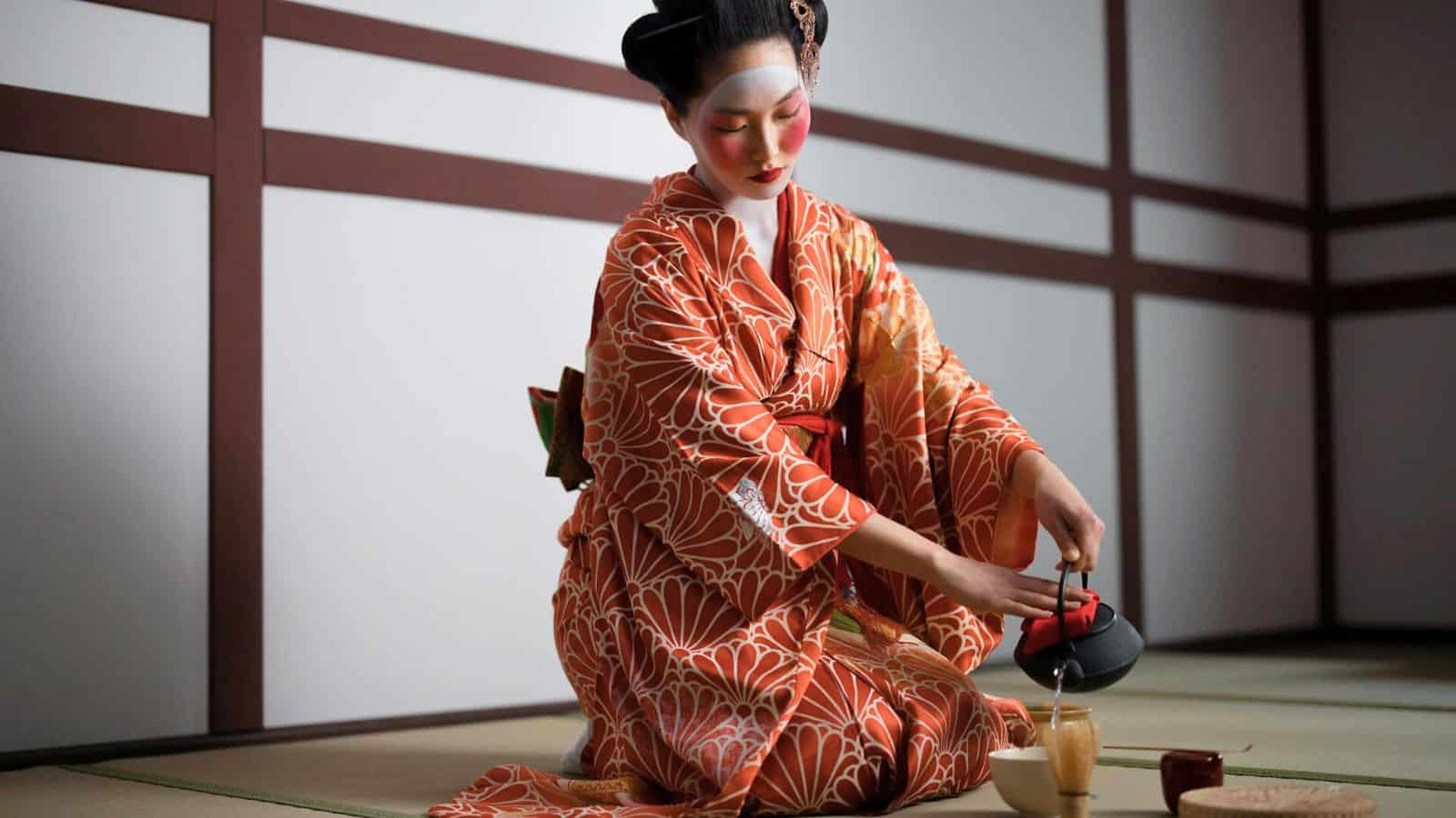A person dressed in a traditional Japanese kimono is performing a tea ceremony. They are kneeling on a tatami mat and carefully pouring tea from a teapot into a cup. The background features a wall with wooden panels.