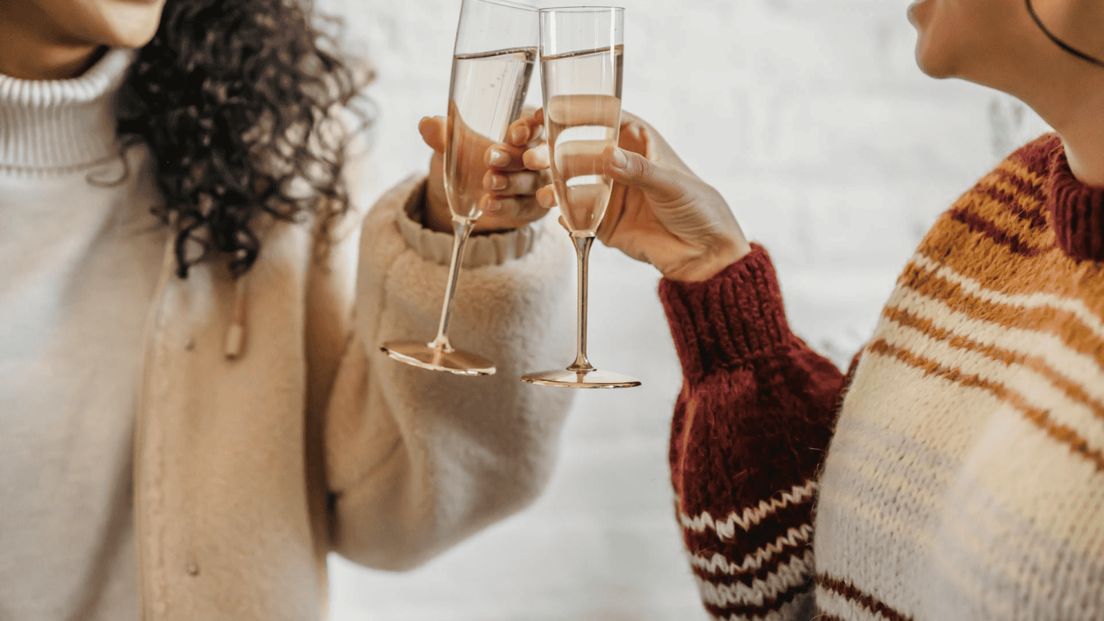 Two people clink champagne glasses together, toasting perhaps to the new harvest of unusual plants for winemaking. One wears a cream sweater, the other a striped sweater. The background is softly out of focus, suggesting an indoor celebratory setting.