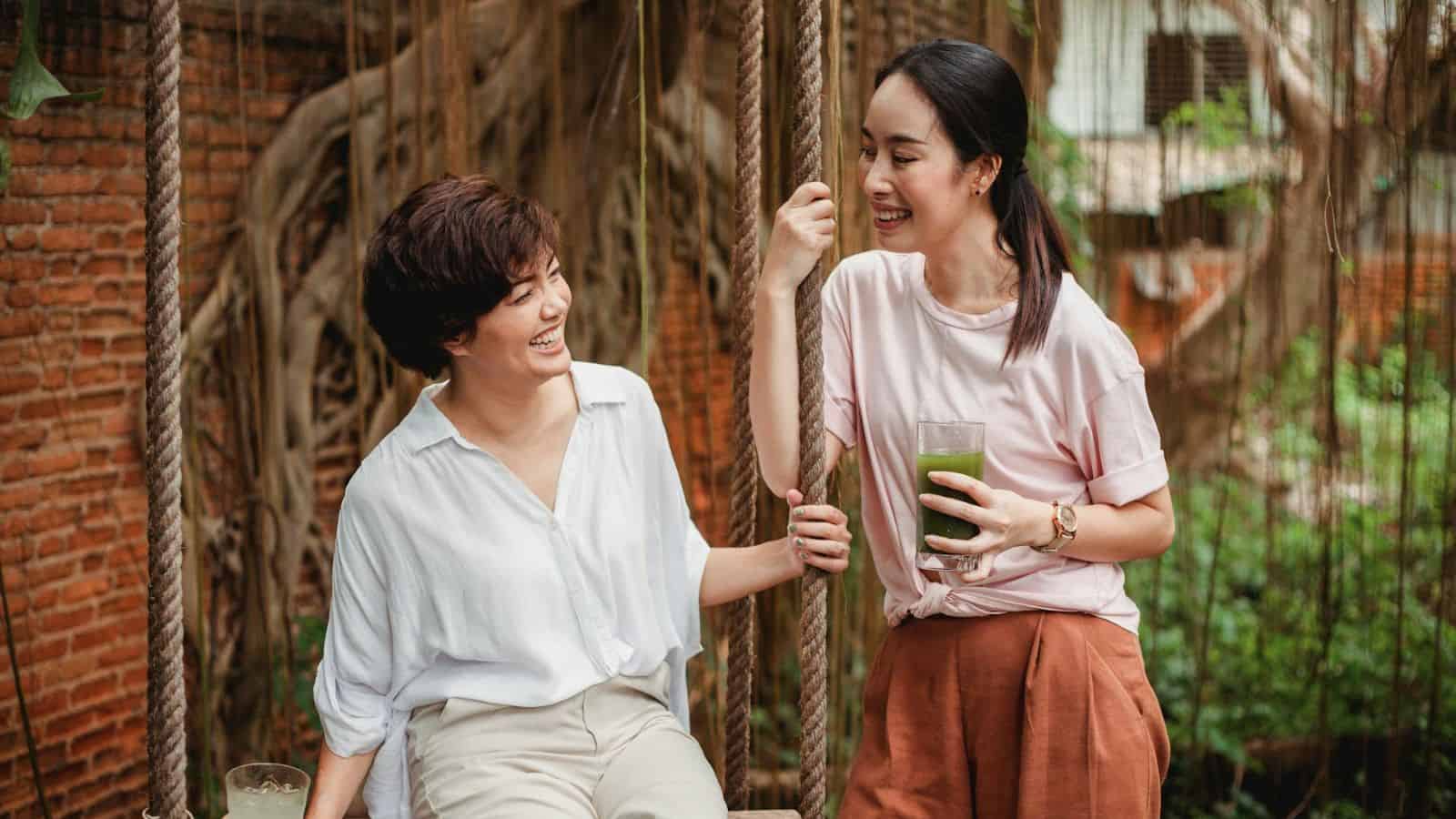 Two women are sitting and standing on a wooden swing. One woman sits, wearing a white shirt and light-colored pants. The other stands, holding a green drink in one hand, and wears a light pink shirt and rust-colored pants. They are smiling and talking.