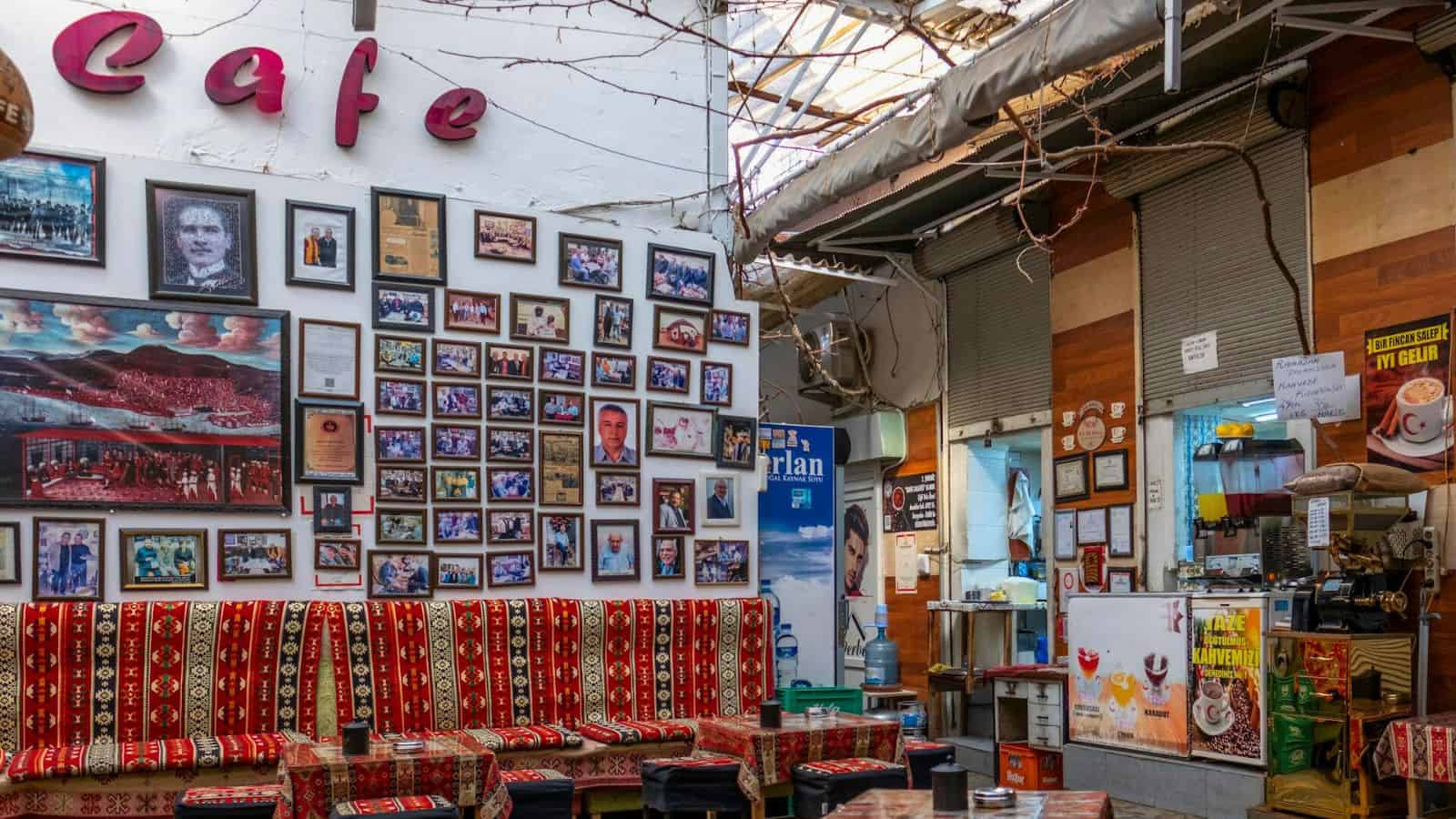 An indoor cafe with colorful patterned cushions on benches, walls adorned with numerous framed photos and memorabilia. A small counter space with various items and a menu board is to the right. The ceiling has exposed beams and partial skylights.