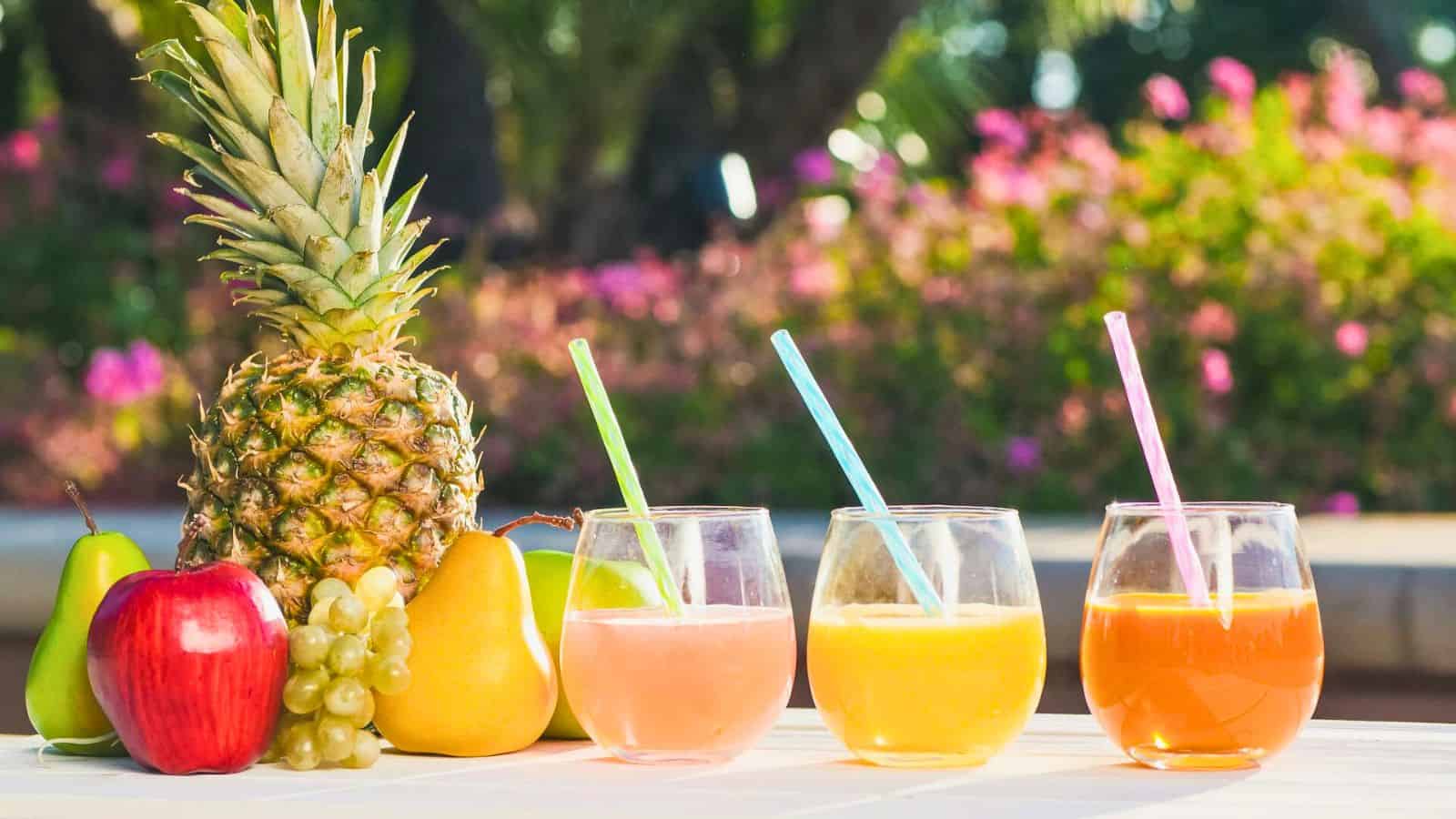 The image shows a pineapple, pear, apple, and bunch of grapes on a table next to three glass cups of juice. The juices are pink, orange, and red, each with a straw. The background is a garden with blurred trees and pink flowers.