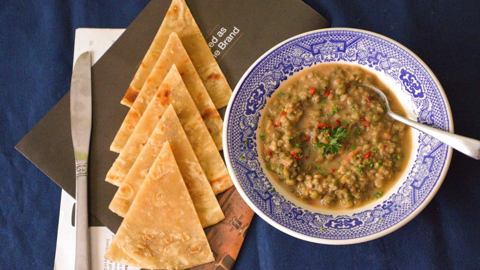 A bowl of lentil soup garnished with chopped herbs and red chili flakes, with a spoon inside, is placed on a blue-patterned ceramic plate. Accompanying the soup are five triangular pieces of flatbread and a knife arranged next to the bowl on top of several papers.