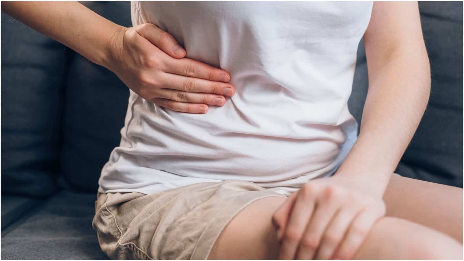 A person wearing a white shirt and beige shorts is sitting on a dark-colored couch with their left hand placed on their lower right abdomen. Their right hand rests on their knee.