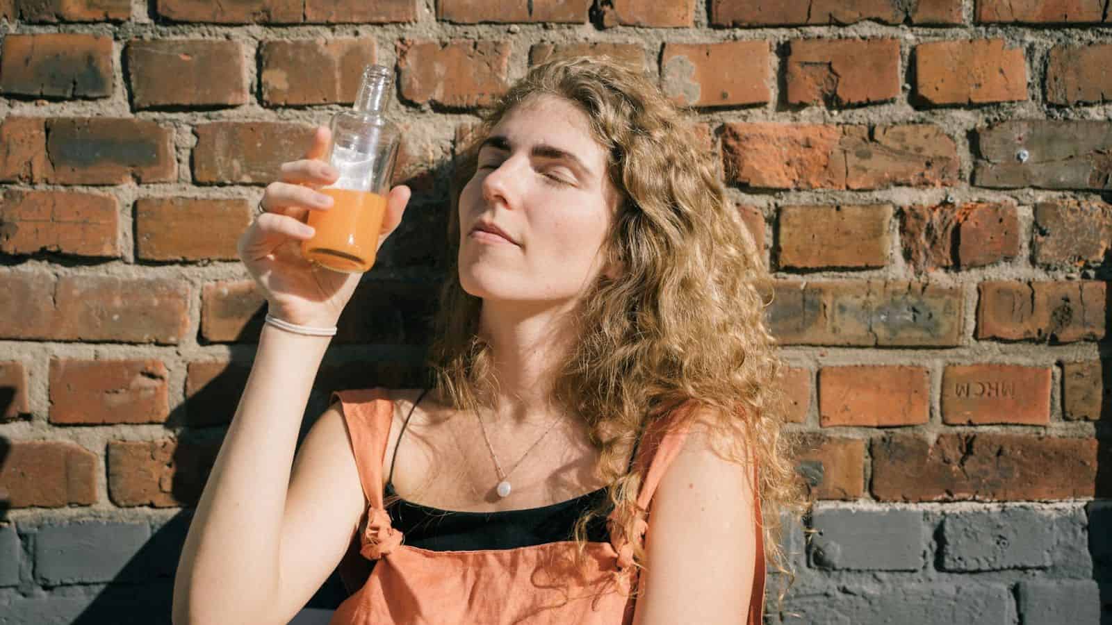 A young woman with curly hair is sitting against a brick wall, holding a bottle of orange-colored liquid up to her face. Her eyes are closed and she appears to be enjoying the sunlight on her face. 