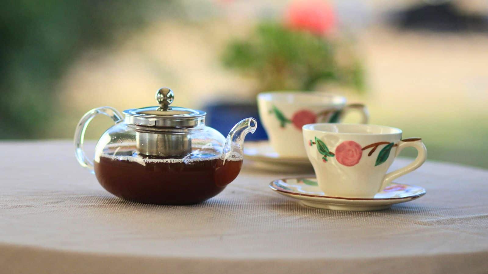 A glass teapot with a metal infuser filled with tea is placed on a tablecloth-covered surface. Two floral-patterned teacups with saucers are positioned behind the teapot. There is a blurred background with greenery and a hint of red.