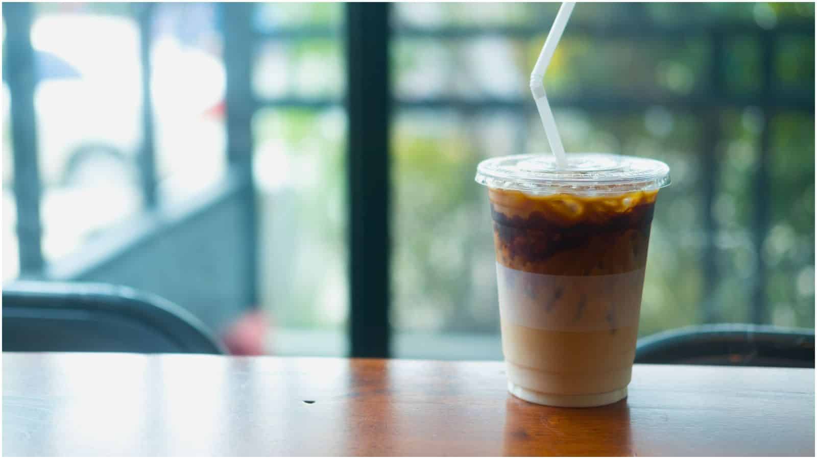 A plastic cup of iced coffee with a clear lid and a white straw sits on a wooden table. The coffee appears layered, with milk on the bottom and a darker coffee layer on top. The background shows a blurred view of windows and greenery outside.