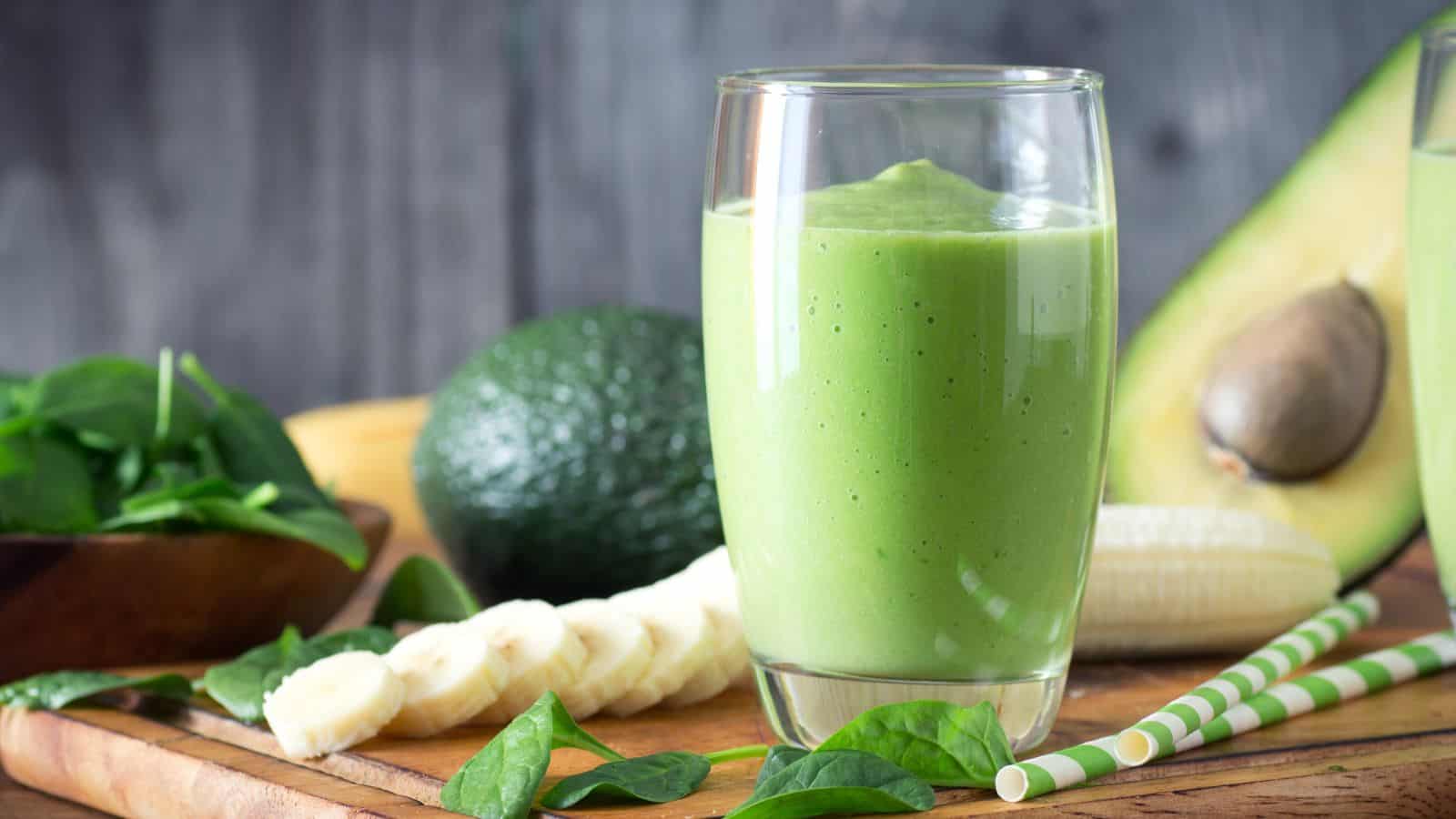 A glass of green smoothie is placed on a wooden surface. Surrounding the glass are fresh ingredients including avocado, banana slices, and spinach leaves.