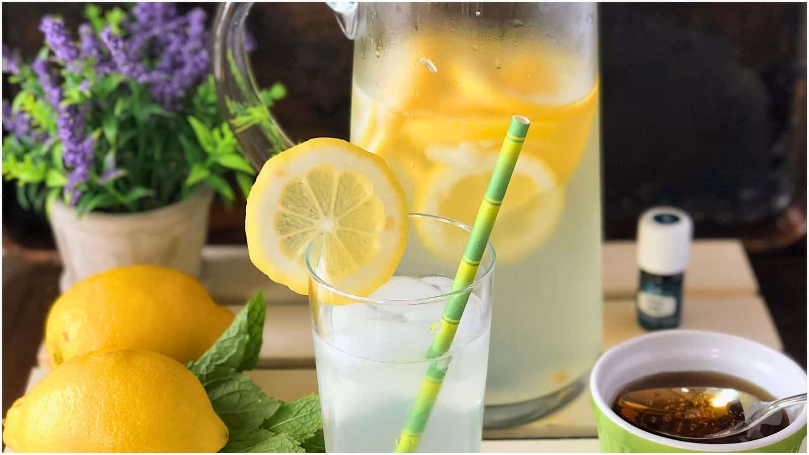 A glass pitcher filled with lemon slices and lemonade stands on a table next to a glass with lemonade and a green straw. Fresh lemons, a sprig of mint, a small bowl of honey, and a potted plant with purple flowers are also on the table.