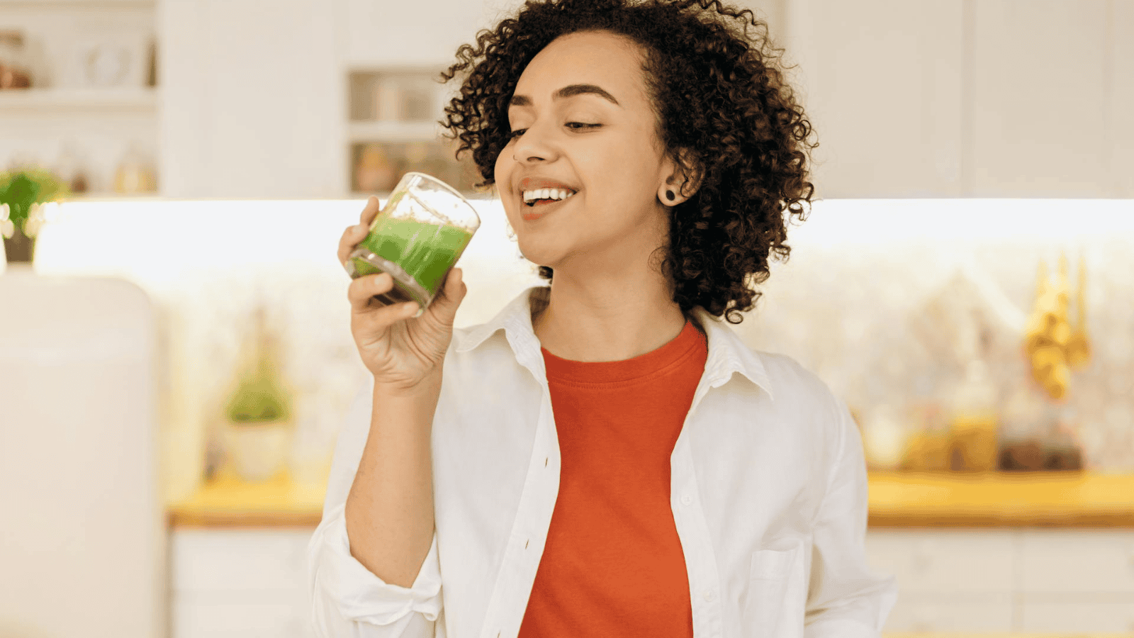 A person with curly hair stands in a sleek, modern kitchen, admiring a glass of superfood smoothie. Clad in a white shirt over a red top, they savor the vibrant green drink, clearly enjoying its nutritious goodness.