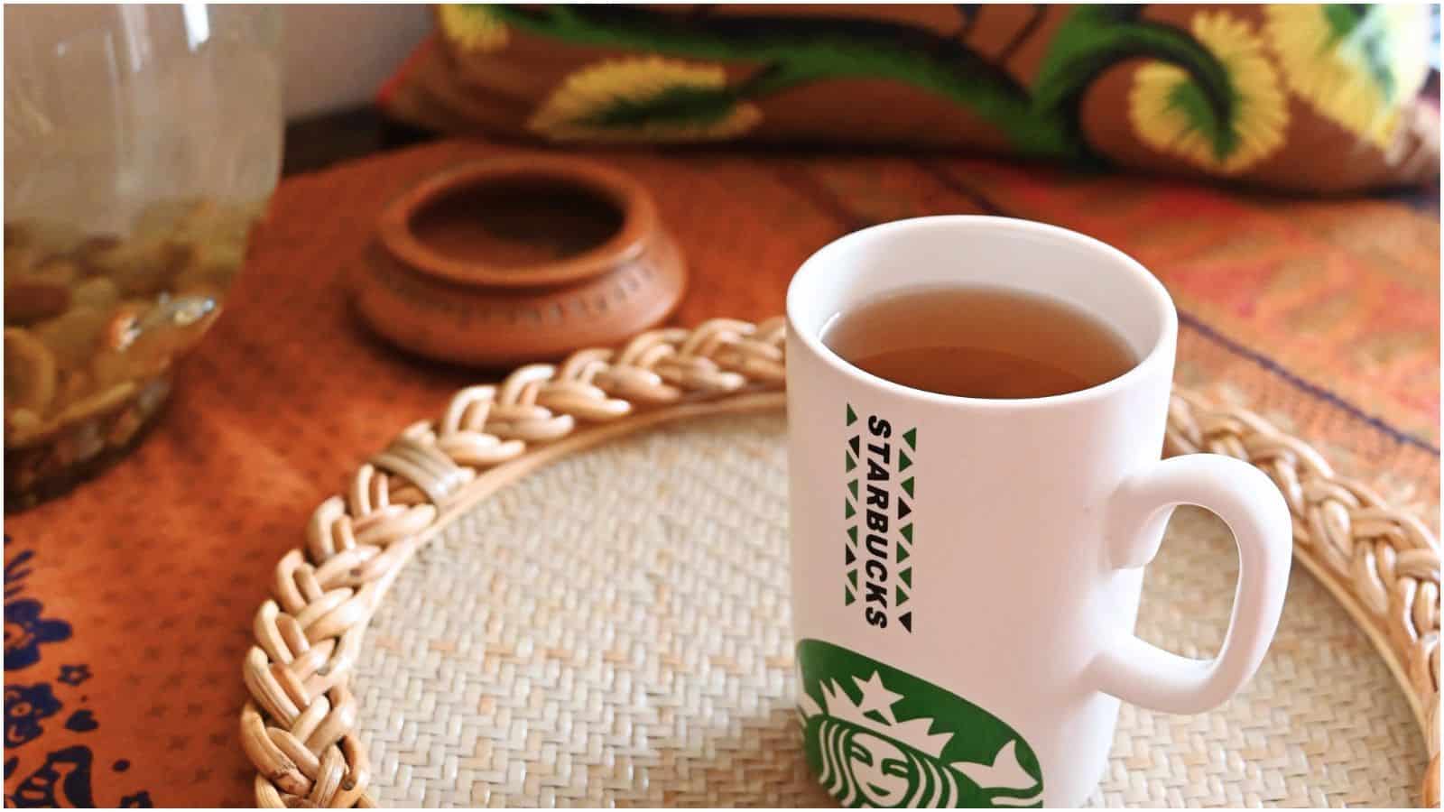 A white Starbucks mug filled with tea is placed on a woven tray atop a textured surface. In the background, there is a glass container with pebbles and a decorative item with a colorful design.