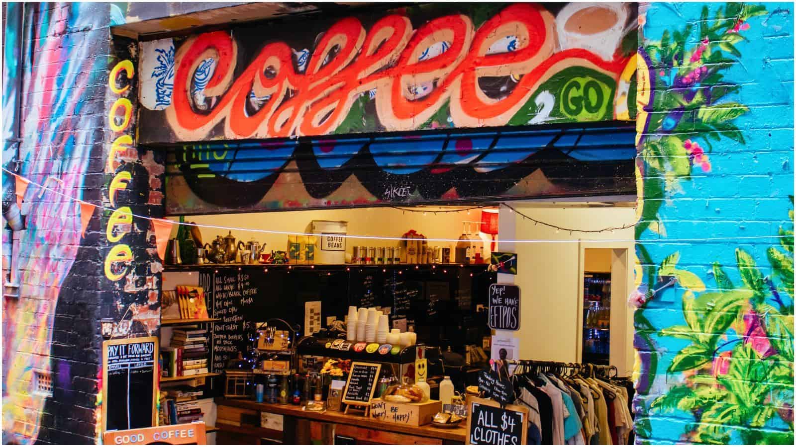 A street-side coffee shop entrance featuring vibrant graffiti art spelling "coffee." Inside, the shop showcases shelves with merchandise, a counter with coffee-making supplies, a chalkboard menu, and racks of clothing.