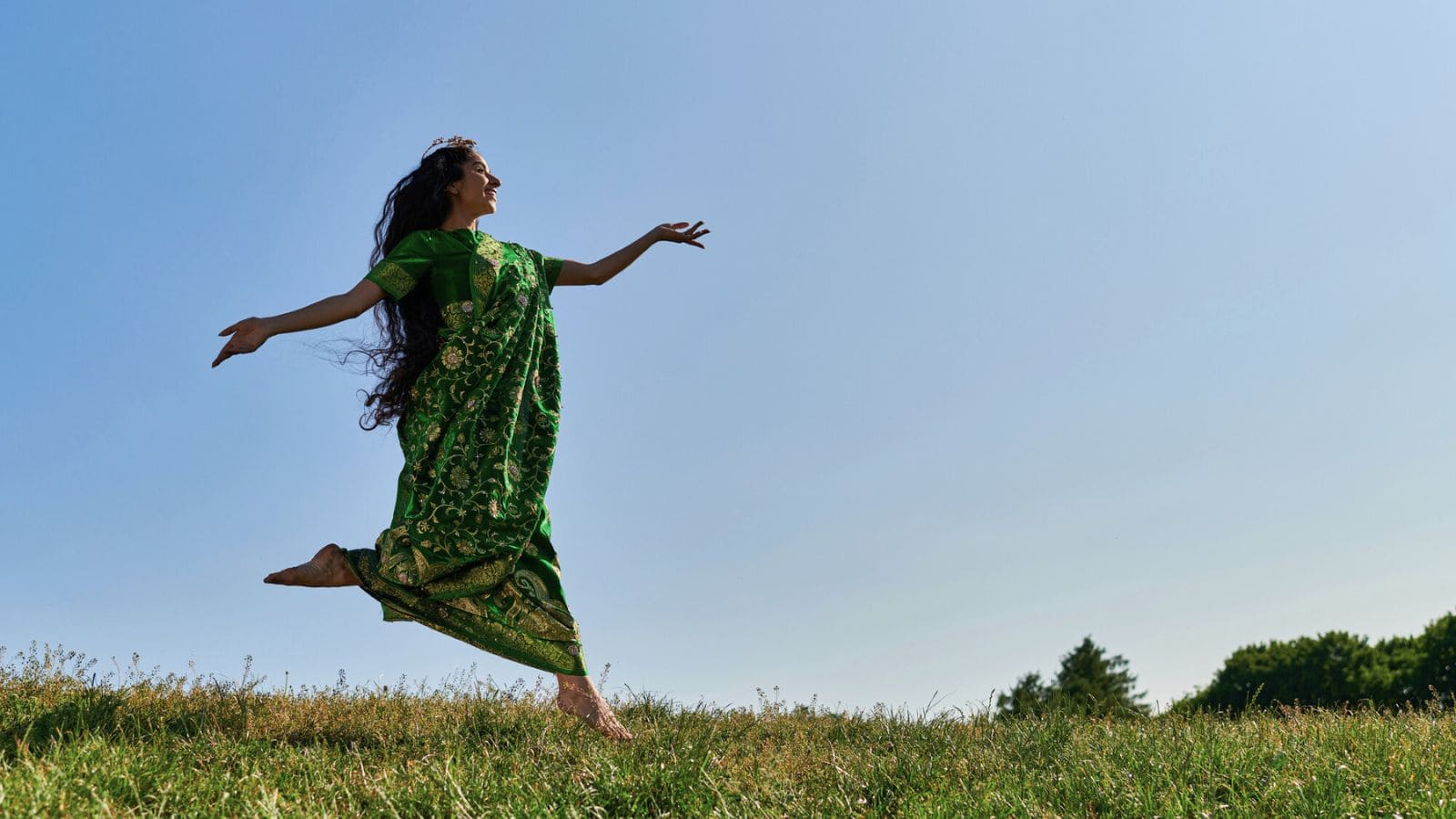 A person wearing a green, gold-embroidered sari is captured mid-jump on a grassy field. With long, flowing hair and arms extended against a clear blue sky, they appear to be joyfully leaping. Trees are visible in the background.