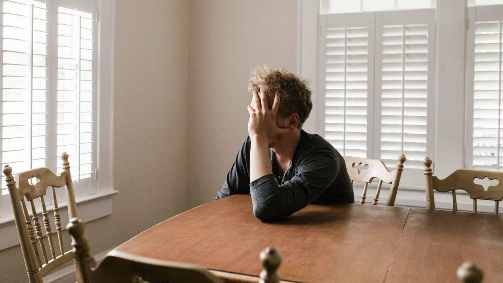 A person with curly hair is sitting at a wooden dining table with their head resting in their hand, looking out the window. The room has white walls and multiple shuttered windows. Five wooden chairs surround the table.