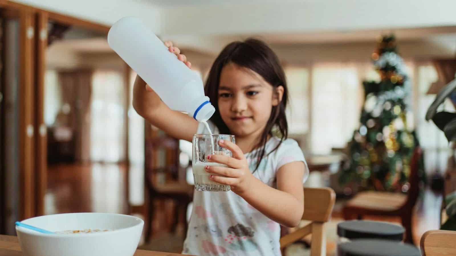 A young girl is pouring milk from a plastic bottle into a glass at a dining table. A bowl with cereal and a spoon is in front of her.