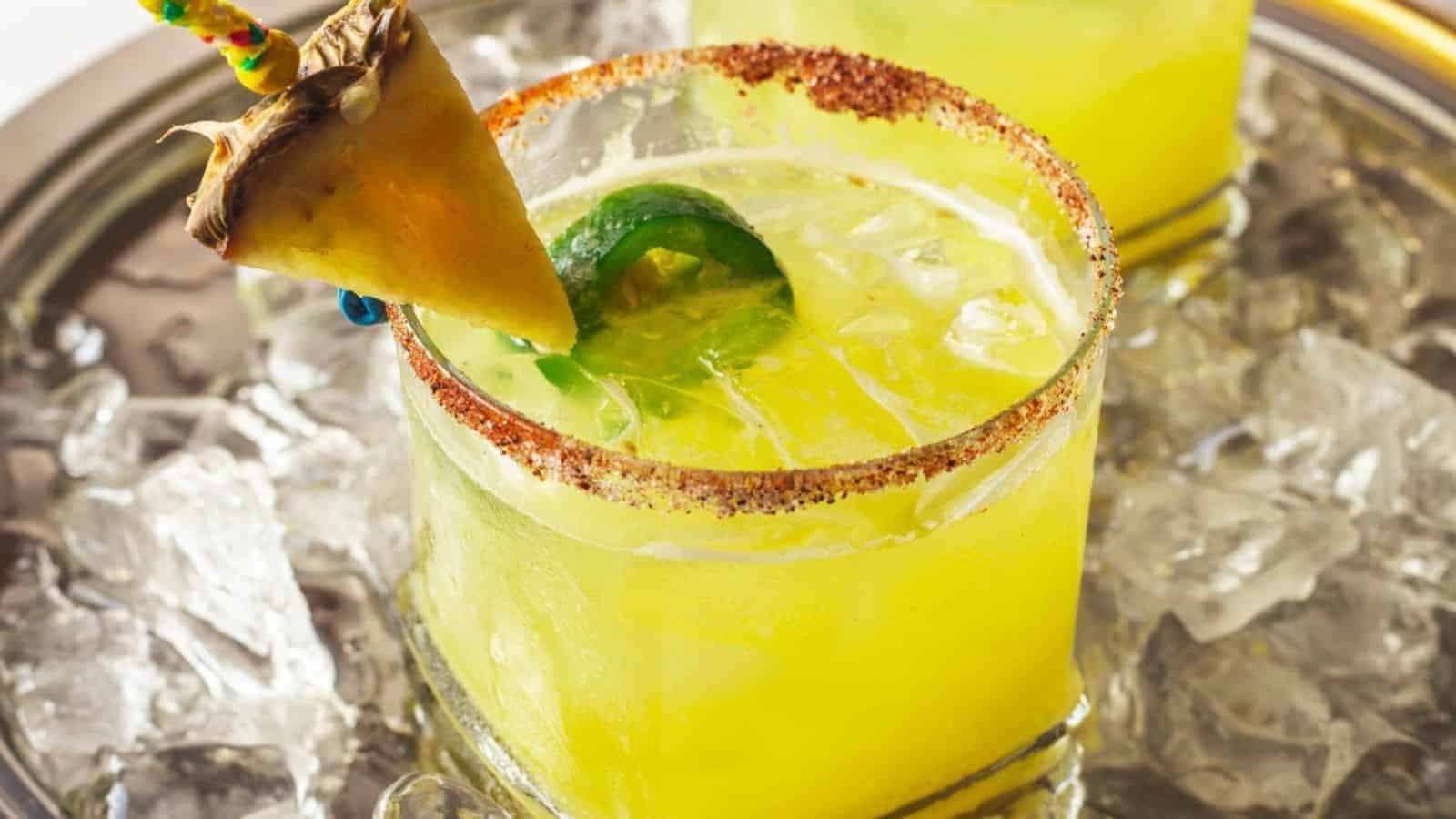 A fun non-alcoholic yellow cocktail in a glass rimmed with chili powder, garnished with a slice of pineapple and a jalape&ntilde;o slice. The glass rests on a tray filled with ice. Another similar drink is partially visible in the background.