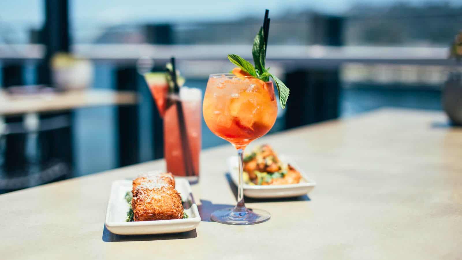 A table displays a bright orange drink with a straw and mint garnish, alongside two rectangular plates of food. One contains a pastry dusted with powdered sugar, and the other features a serving of meat topped with garnish. Another fun bar order can be seen in the background.