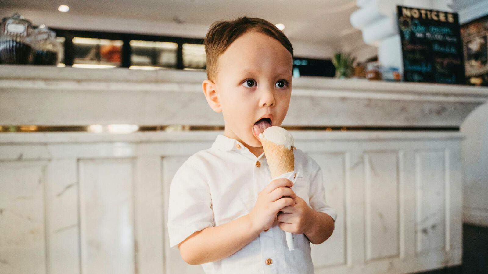 A young child with short brown hair is standing inside a well-lit room, wearing a white shirt, and licking an ice cream cone. The background features a white counter with a chalkboard menu on the right side.