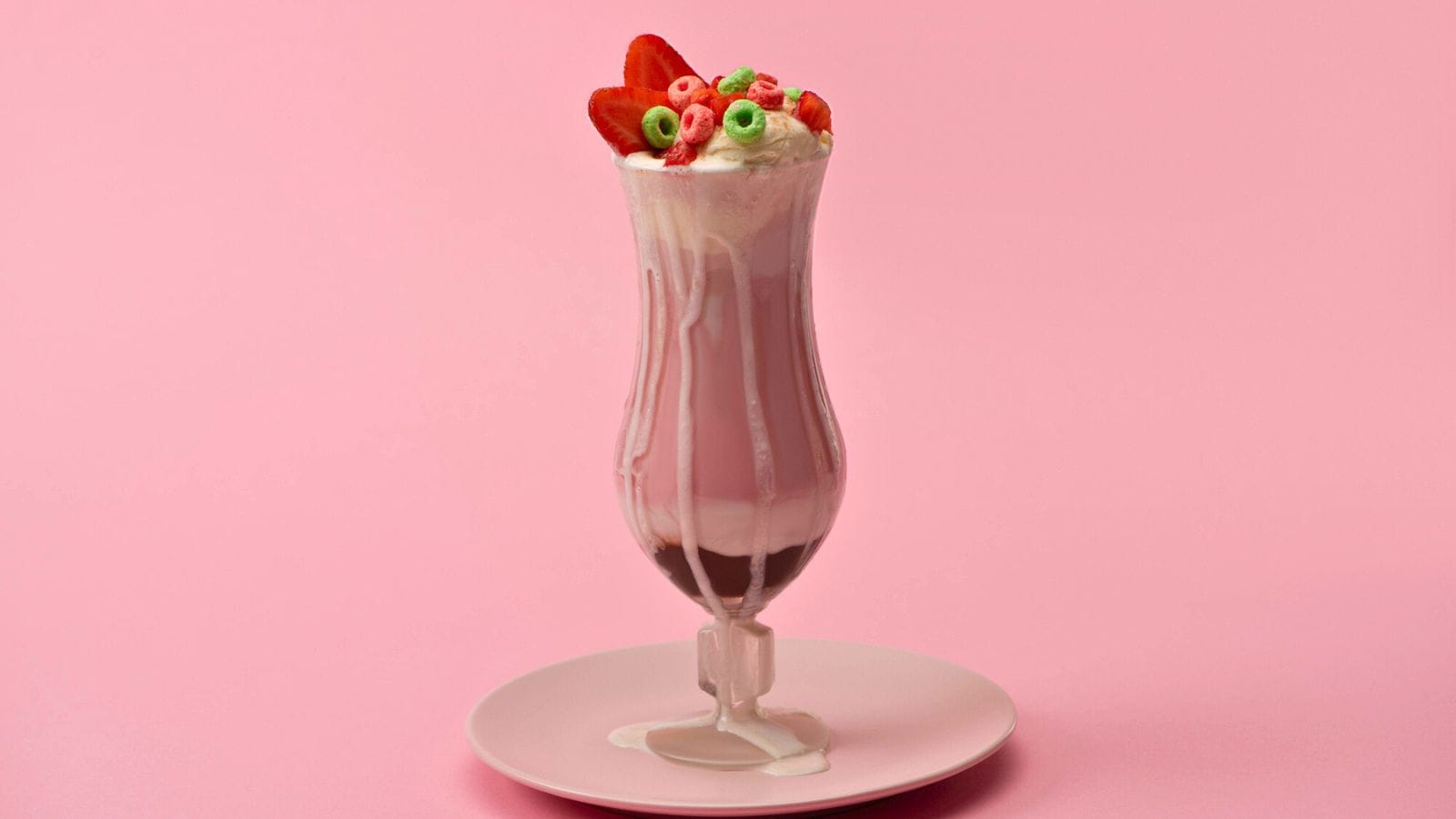 A strawberry milkshake in a tall glass with whipped cream, sliced strawberries, and colorful cereal pieces, placed on a plate. The pink background complements the color of the milkshake and the toppings. 