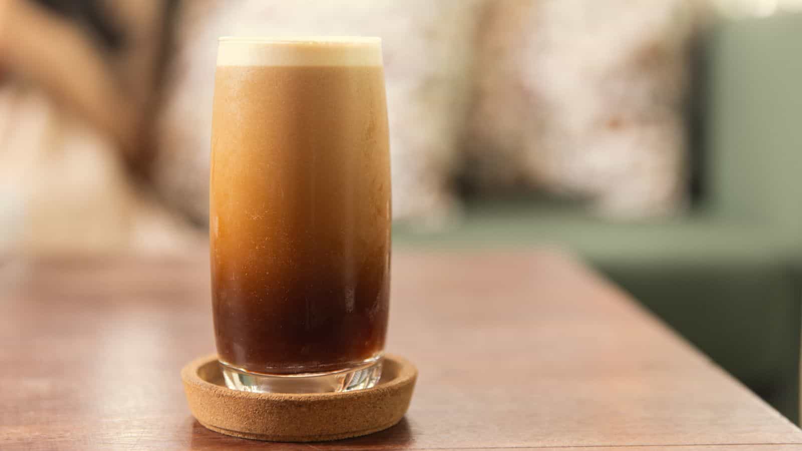 A tall glass filled with a layered iced coffee sits on a wooden coaster. The top layer is a frothy, light-colored foam, while the bottom layer is a dark coffee. The glass rests on a wooden table with a blurred background.