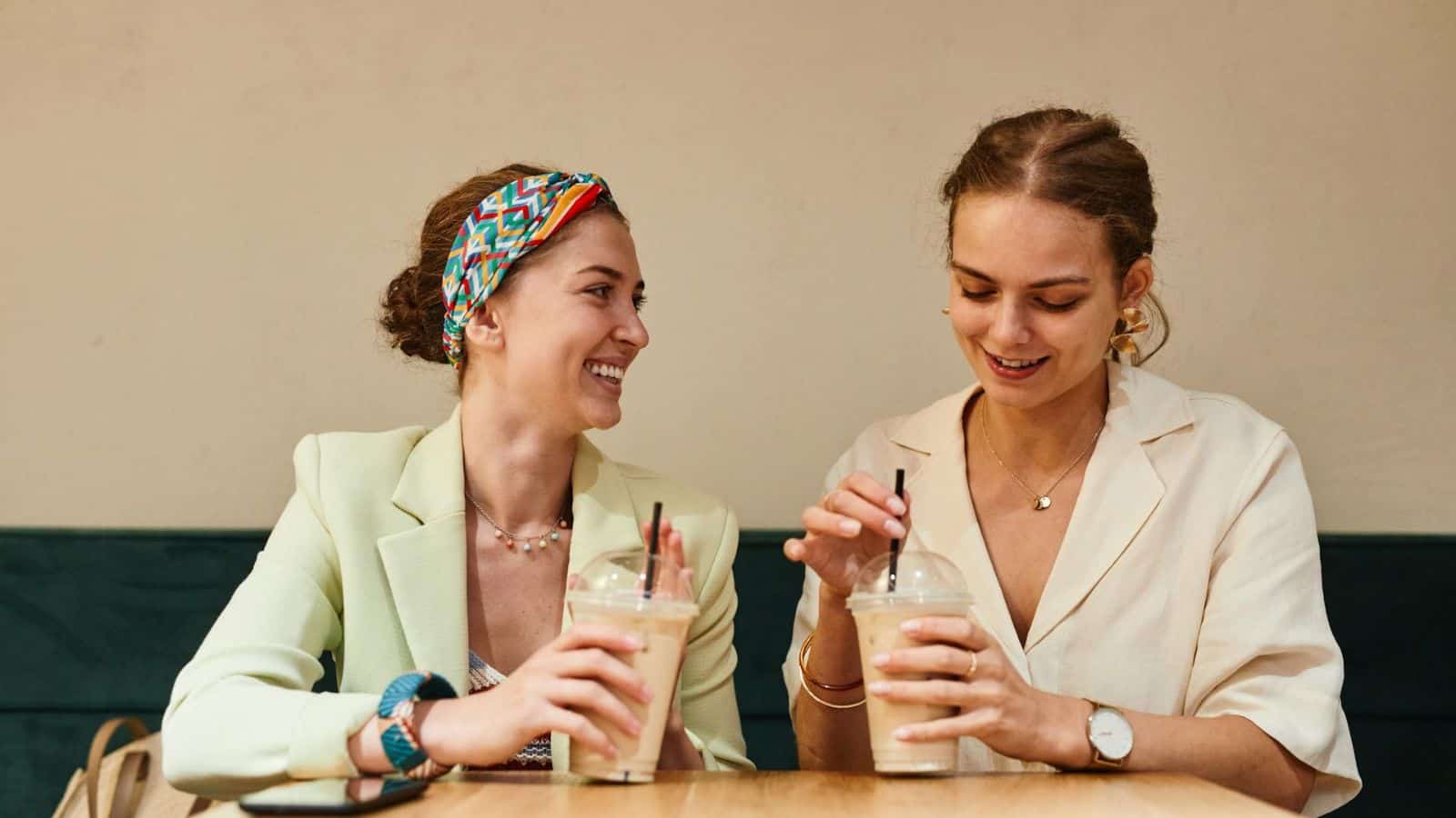 Two women are sitting at a table, each holding a cup with a straw. The woman on the left is smiling and wearing a colorful headband, while the woman on the right looks down at her drink and wears a wristwatch. 