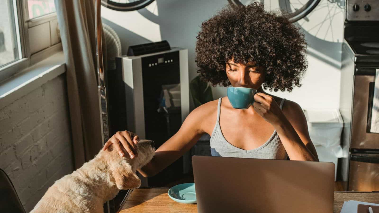 A woman sipping a cup of some beverage and patting her dog on the head