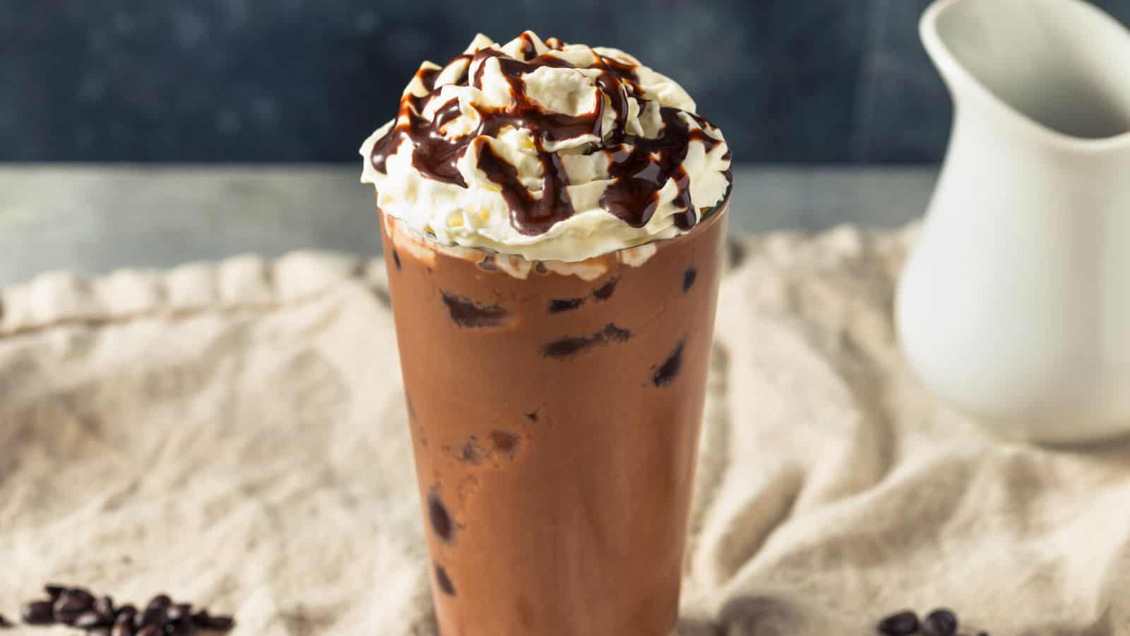 A tall glass of iced coffee topped with whipped cream and chocolate drizzle is placed on a cloth-covered surface. A small white pitcher sits in the background.