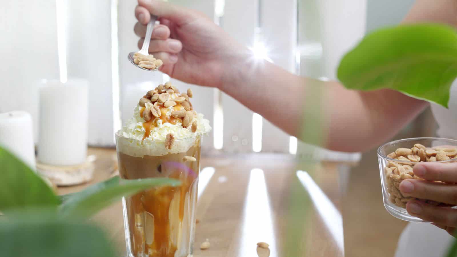 A person is holding a spoonful of peanuts over a glass of iced coffee topped with whipped cream, caramel drizzle, and peanuts. The person also holds a small cup of peanuts while standing in a bright, airy room with green plants in the foreground.