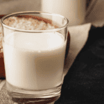 A glass of oat milk with a bowl of oats in the background