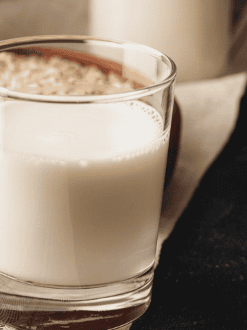 A glass of oat milk with a bowl of oats in the background