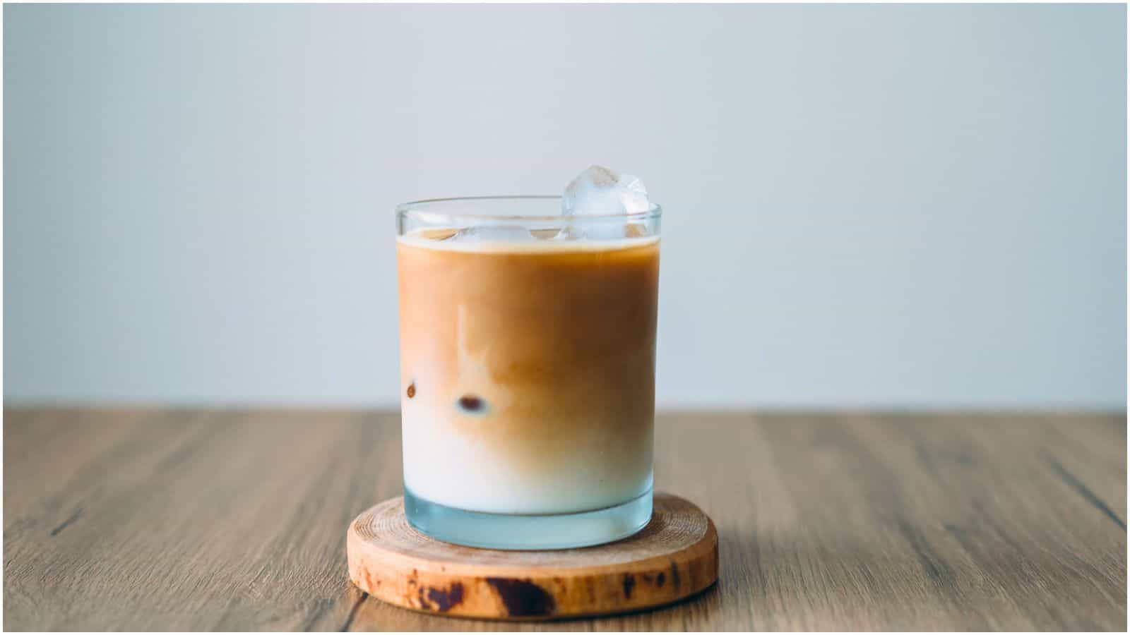 A glass of iced coffee with milk is placed on a wooden coaster. The drink has a visible layer separation, with darker coffee on top and lighter milk below. Three ice cubes float in the coffee. The background is plain and light-colored.