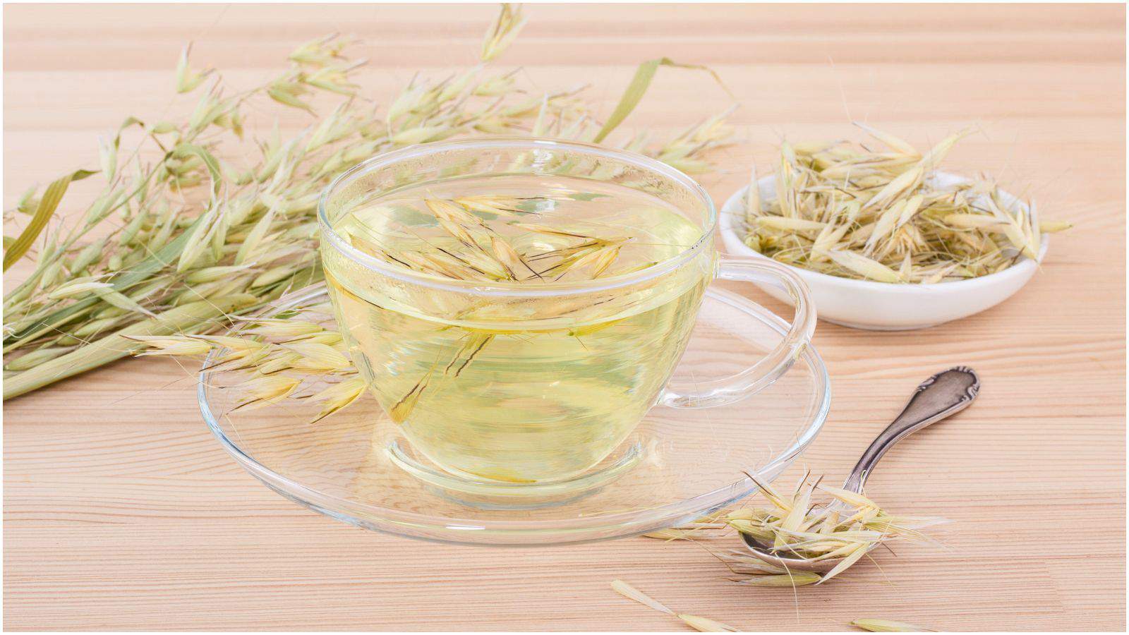 A glass cup filled with a light greenish-yellow herbal tea is garnished with oat stems and sits on a clear saucer on a wooden surface. Nearby, additional oat stems are scattered around, some lying next to a silver spoon and a small white bowl filled with oats.