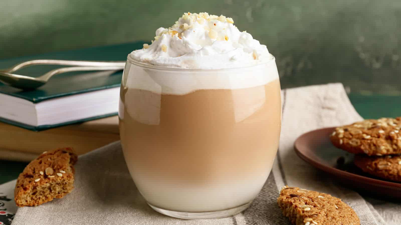 A frothy latte topped with a dollop of whipped cream and sprinkled with nuts in a clear glass sits on a beige napkin. Next to it are a stack of books, a spoon, and a plate with several sesame cookies.
