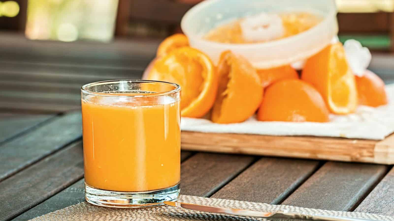 A glass of orange juice is on a wooden table with a serrated knife on a placemat in the foreground. In the background, there is a cutting board with several halved and peeled oranges next to a plastic container.