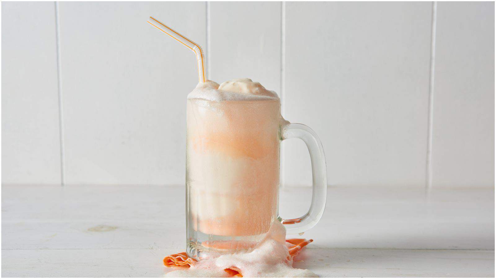 A glass mug filled with an orange and white ice cream float is placed on a light-colored surface against a white tiled background. The float is foaming over the rim with a straw in the mug and an orange napkin underneath.