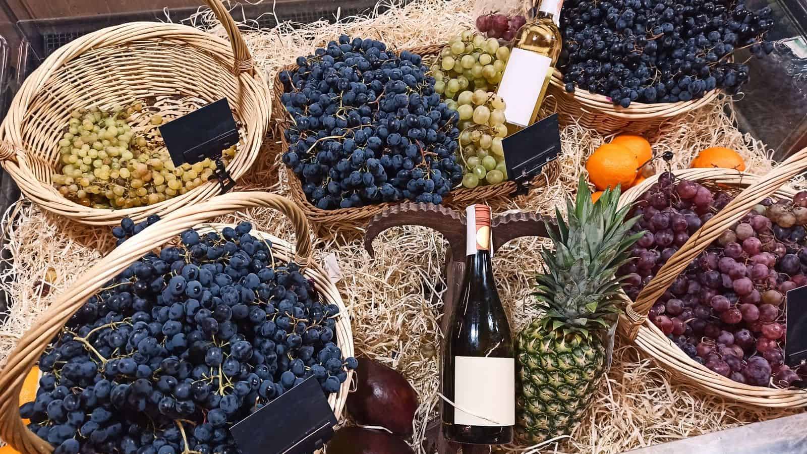 Baskets of green and purple grapes, a pineapple, two bottles of wine, oranges, and a few dark vegetables are arranged on a bed of straw. Blank labels are displayed on some baskets.