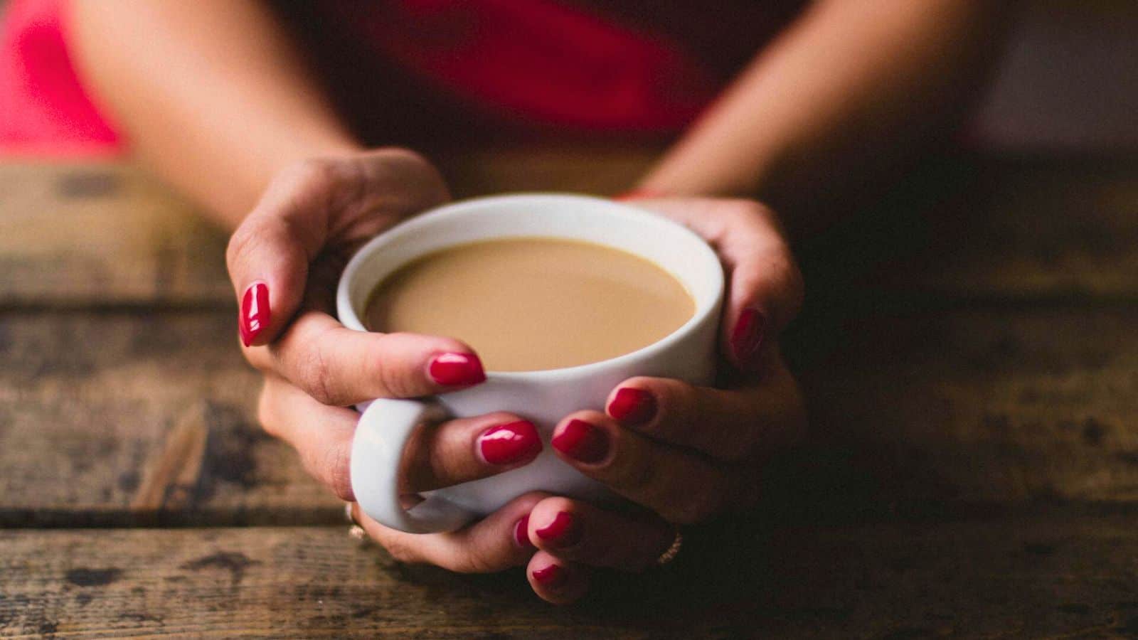 A person with red nail polish holds a white mug filled with a light brown beverage. The hands are resting on a rustic wooden surface.