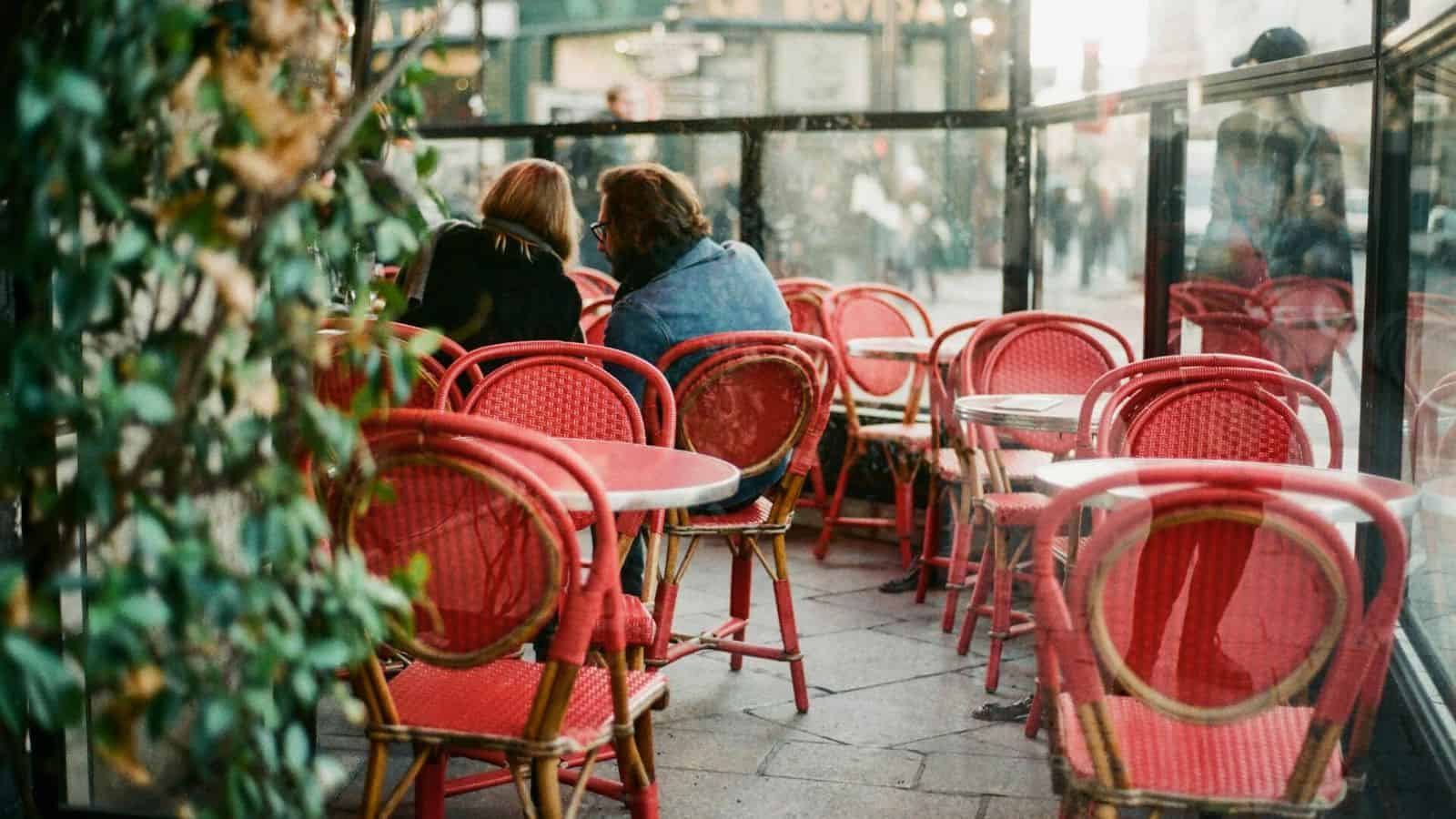 Two people are sitting at a table with red chairs in a partially enclosed outdoor caf&eacute;. The chairs have a woven pattern. The background shows blurred images of the street and people walking, with green plants visible on the left side of the image.