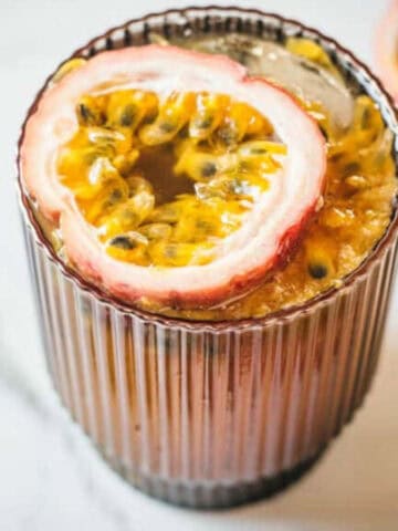 A brown ribbed glass containing a passion fruit beverage with half a passion fruit resting on top. Another half passion fruit is in the background on a white marble surface. The drink appears to have seeds and pulp floating in it.