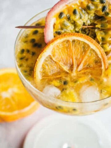 A cocktail in a glass filled with ice, topped with passion fruit pulp, and garnished with a slice of passion fruit and a dried orange slice on a skewer. A halved orange sits in the background on a marble surface.