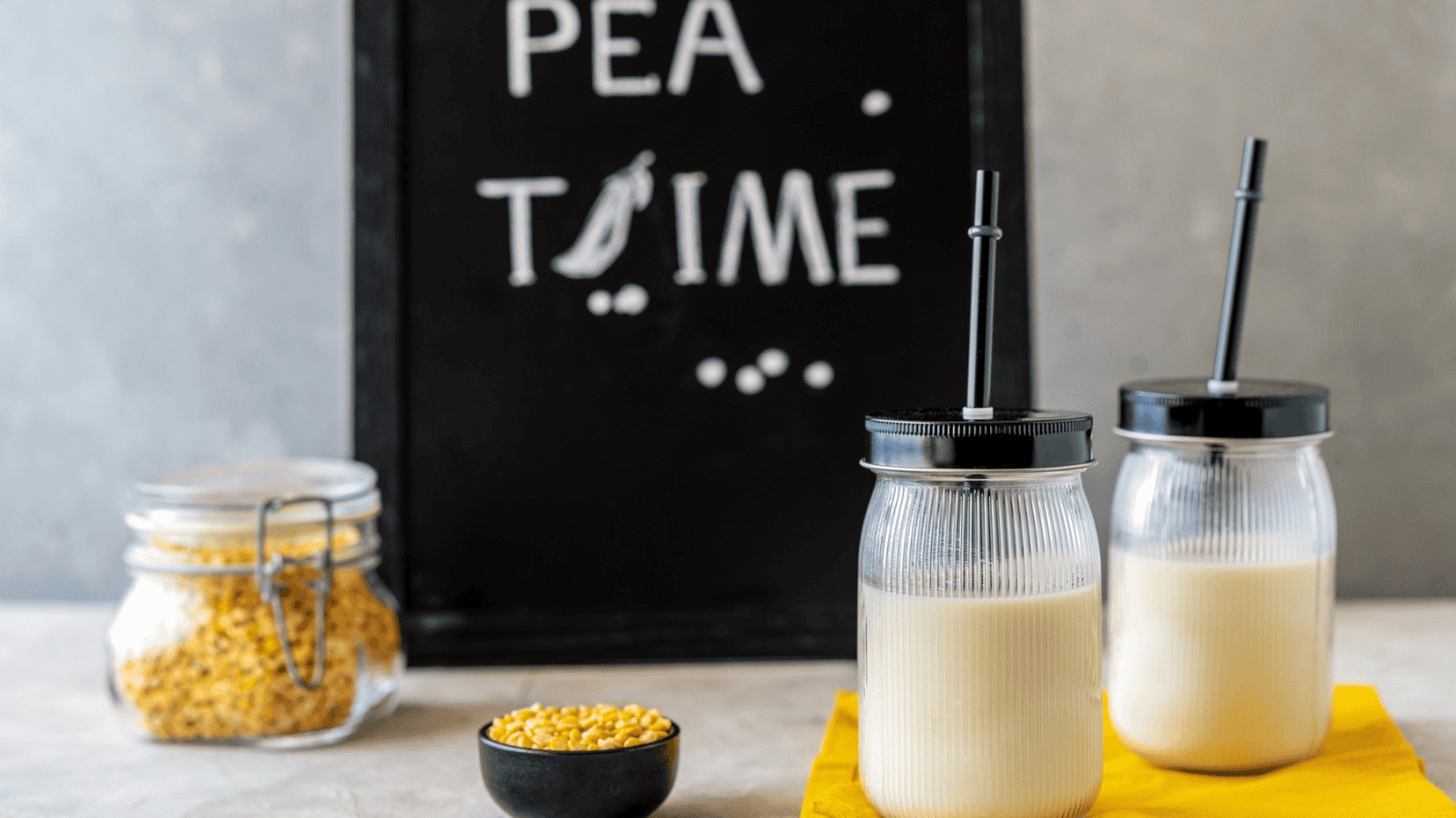 Two glass jars of plant-based dairy milk substitutes, each with a black straw, sit on a yellow cloth. Nearby, a small black bowl of split peas and a glass jar filled with more peas accompany the scene. In the background, a blackboard cheerfully declares "PEA TIME" alongside a pea graphic.