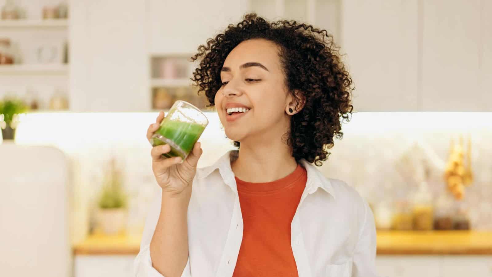 A person with curly hair is standing in a kitchen, holding a small glass of green juice in their left hand and smiling. They are wearing a red shirt with an unbuttoned white shirt over it. The background consists of white cabinets, a backsplash, and some kitchen items.