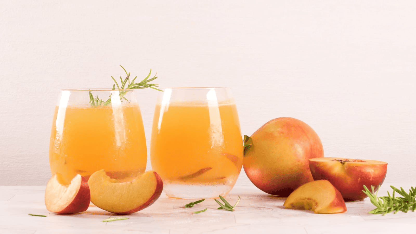 Two glasses of peach juice, garnished with rosemary, sit elegantly on the table. Beside them, whole and sliced peaches are artfully arranged against a white background, hinting at the exotic flavors that unusual plants for winemaking might offer.