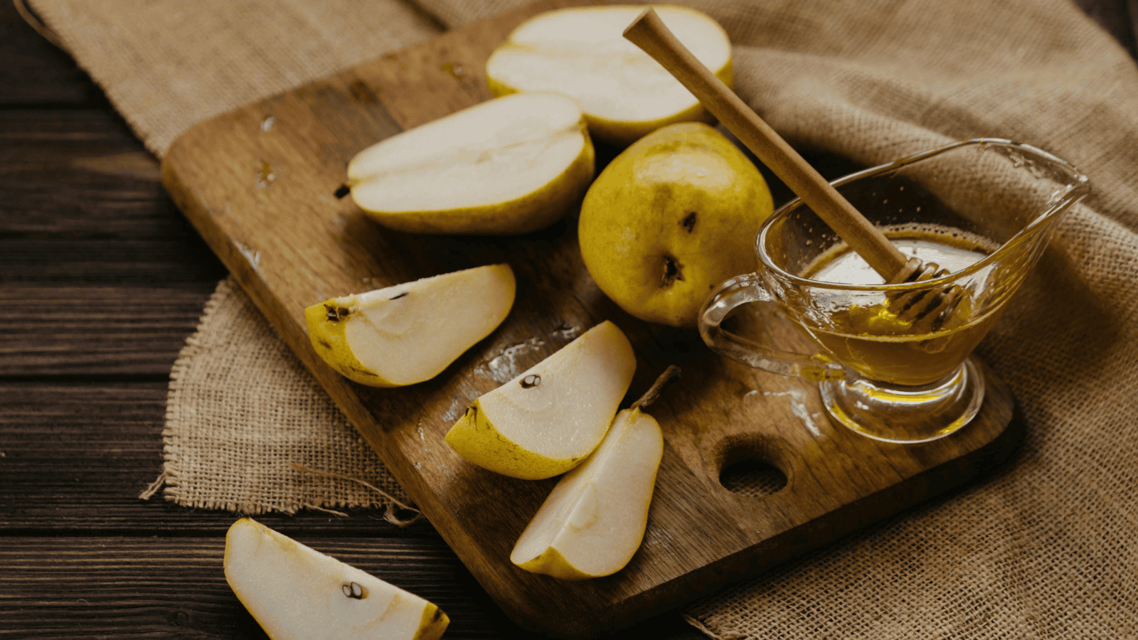 Sliced pears are artfully arranged on a wooden cutting board, accompanied by a glass container of honey infused with cinnamon. The display rests on burlap over a dark surface, echoing the rustic charm often seen in crafting unusual plants for winemaking.
