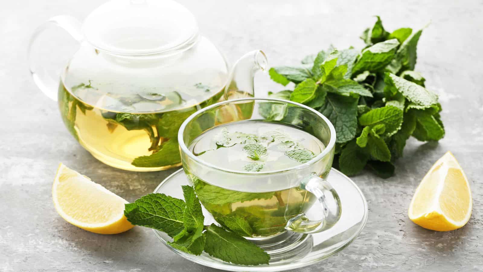 A glass teapot and cup filled with peppermint tea are placed on a grey surface.