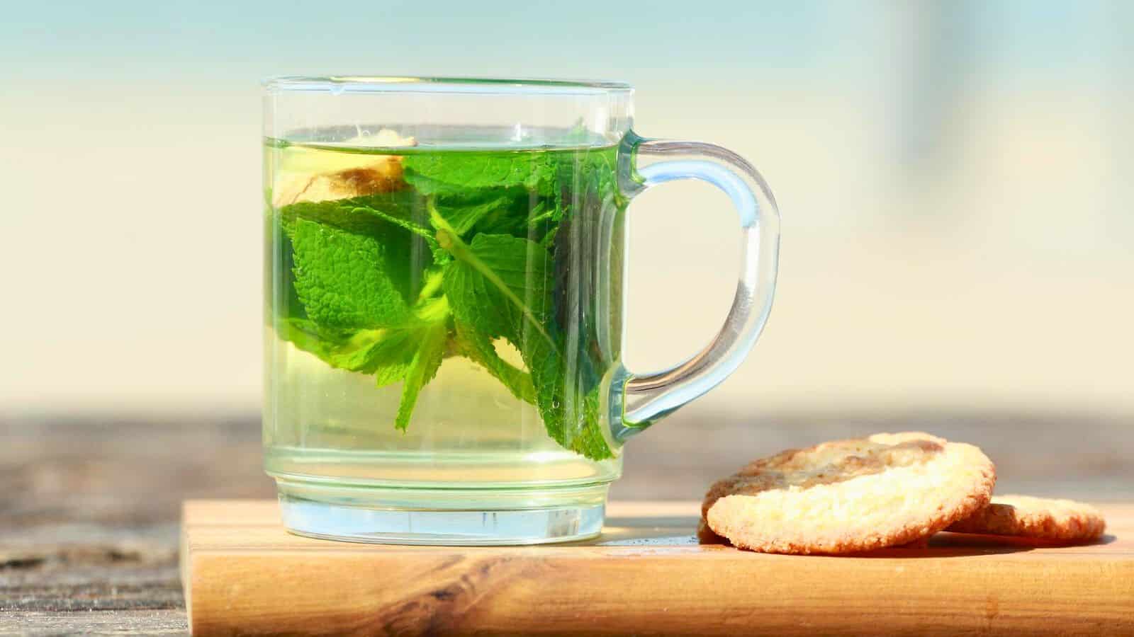 A glass mug filled with herbal tea, garnished with fresh mint leaves and a lemon slice, sitting on a wooden board. Beside the mug, two cookies are placed. 