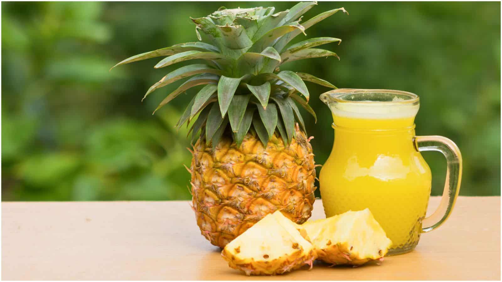A whole pineapple, a glass pitcher filled with pineapple juice, and two pineapple slices are on a wooden surface with a green blurred background.