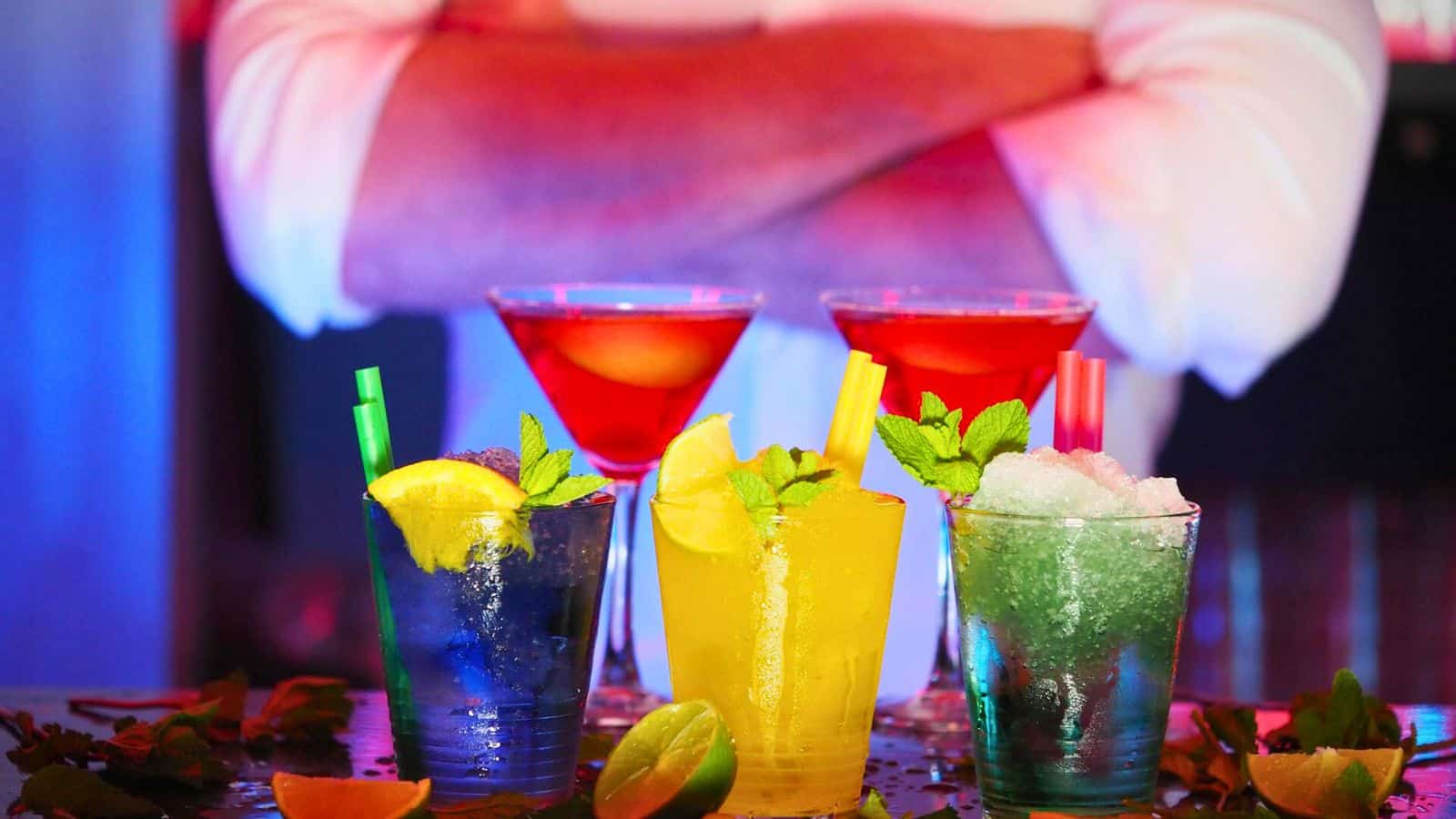 A person with folded arms stands behind a table at a bar, showcasing five colorful cocktails. A blue drink with lemon slice and green straw, a yellow drink with lemon slice and green straw, a green drink with mint leaves and yellow straw, and two red drinks in martini glasses.