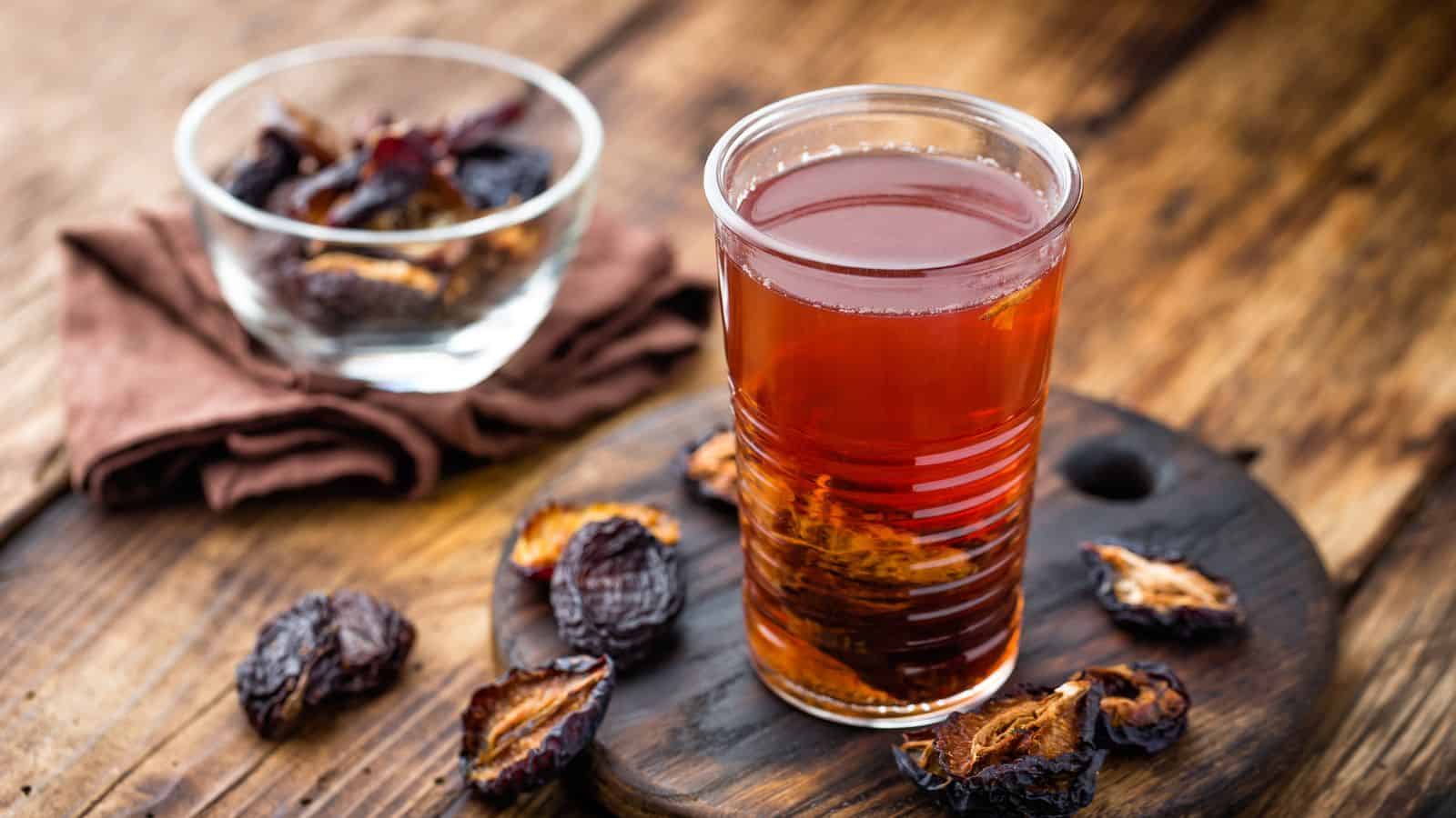 A glass of prune juice is placed on a wooden surface with a few prunes scattered around it. There is a clear bowl filled with more prunes in the background. A folded brown cloth is underneath the bowl. The background is a rustic wooden table.