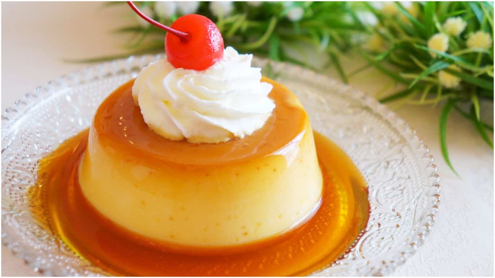 A caramel flan on a clear glass plate. The flan is topped with a dollop of whipped cream and a maraschino cherry. The background contains green foliage and small white flowers.